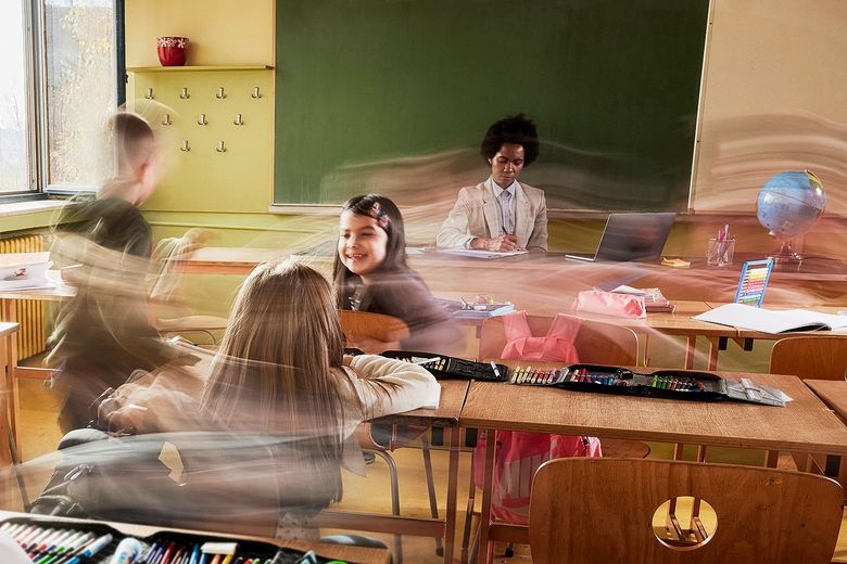 1465764720 In einem Klassenzimmer sitzt eine Frau an einem Schreibtisch vor einer Wandtafel. Vor dem Schreibtisch stehen Schulbänke, zwischen denen drei Kinder stehen. Ihre Bewegungen sind verzerrt dargestellt, was den Eindruck von viel Aktivität und Geschwindigkeit vermittelt.