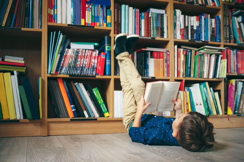 836598276 In einer Bibliothek liegt ein Kind lesend auf dem Rücken und lehnt seine Beine ans Bücherregal.