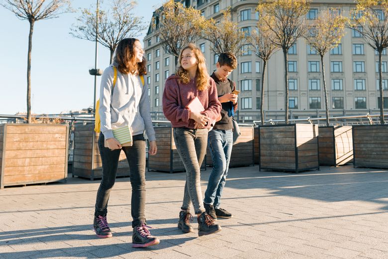 Outdoor portrait of teenage students with backpacks walking and talking. City background, golden hour Drei Jugendliche schlendern über einen Platz.