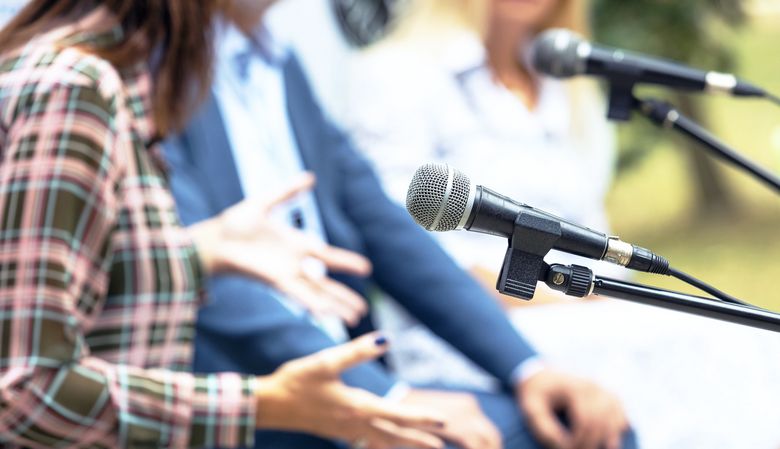 Ein Mikrofon und gestikulierende Hände im Hintergrund deuten eine Diskussion an.