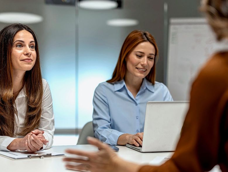 Zwei Frauen sitzen einer dritten Person am Tisch gegenüber.