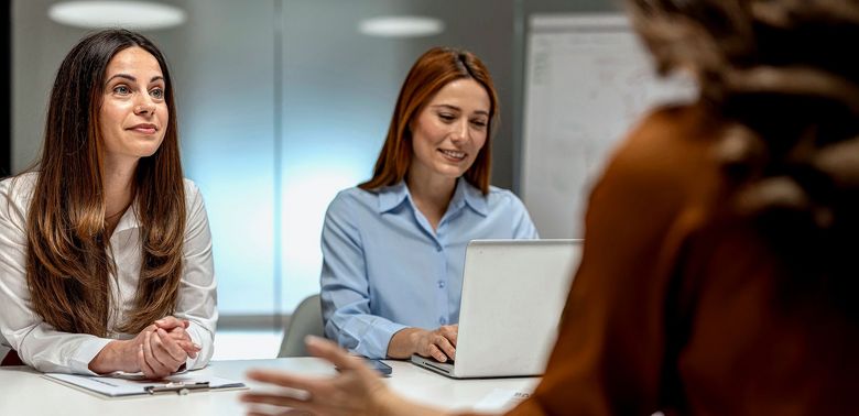 Zwei Frauen sitzen einer dritten Person am Tisch gegenüber.