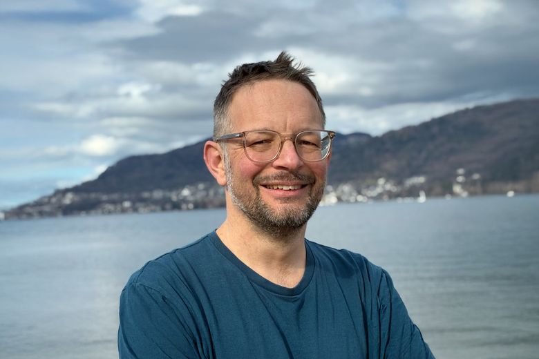 Ein Mann mit Brille steht vor einem See.