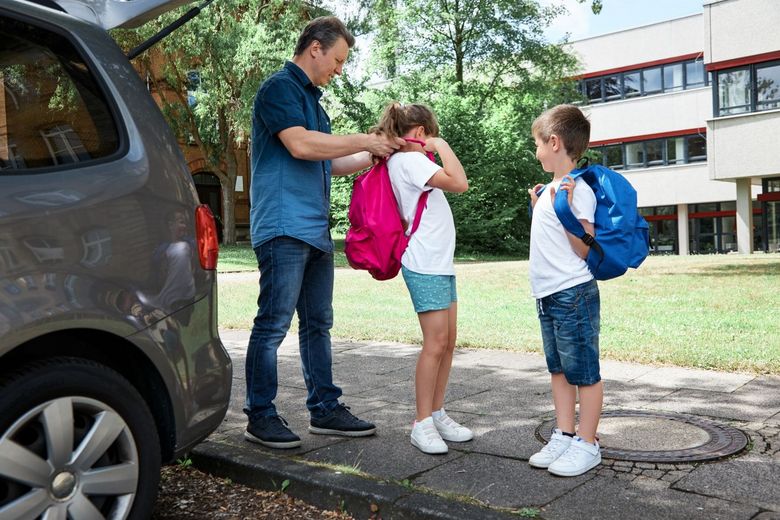 Auto vor einem Schulhaus Vater hilft Tochter beim Anziehen eines Schulrucksacks Sohn schaut zu