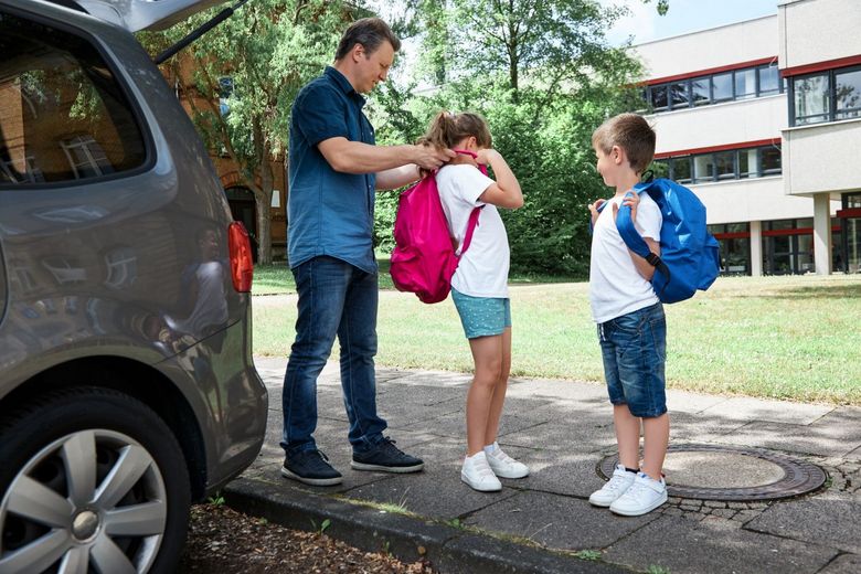 Auto vor einem Schulhaus Vater hilft Tochter beim Anziehen eines Schulrucksacks Sohn schaut zu